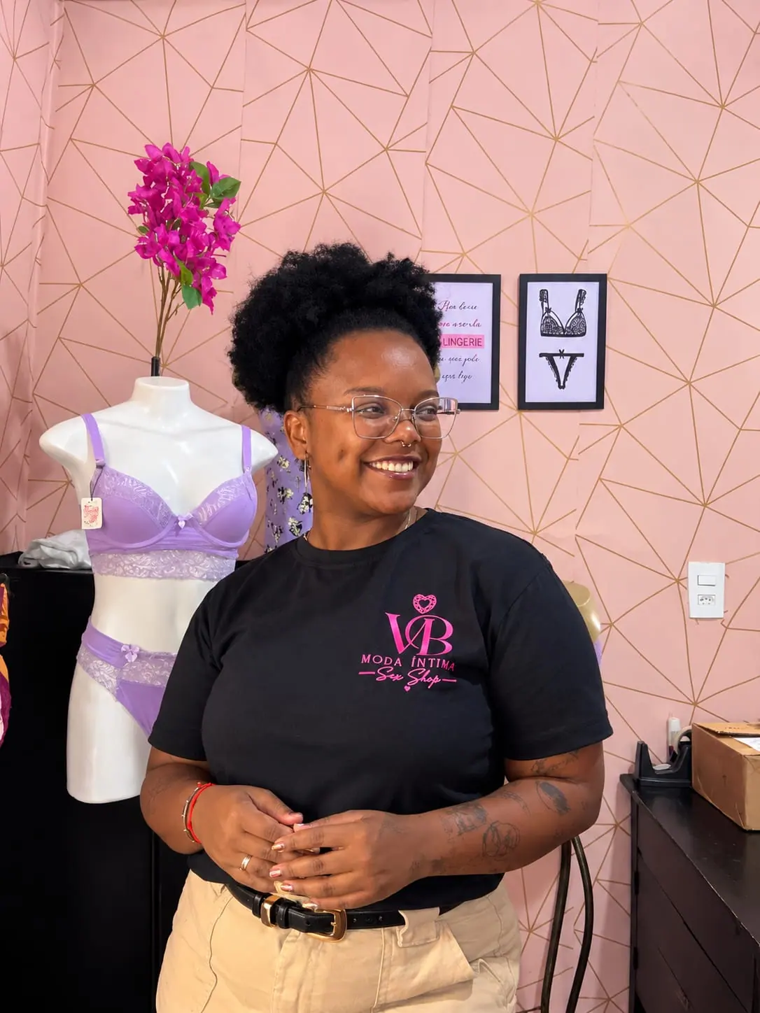 Mulher sorrindo na loja V.B, usando camiseta com a marca, ao lado de um manequim com lingerie lilás.